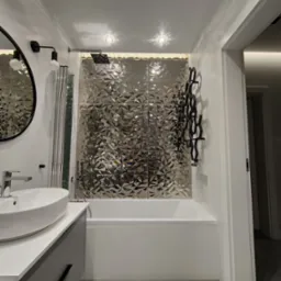 Nowoczesna łazienka z błyszczącymi, srebrnymi płytkami 3D za wanną, designerski grzejnik drabinkowy w kolorze czarnym, okrągłe lustro i biała umywalka nablatowa na szarej szafce.