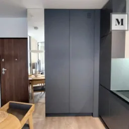 Nowoczesna zabudowa kuchenna w odcieniach szarości z minimalistycznymi frontami, wkomponowana w przestrzeń mieszkania z widocznym fragmentem stołu i krzesła w odbiciu lustra.