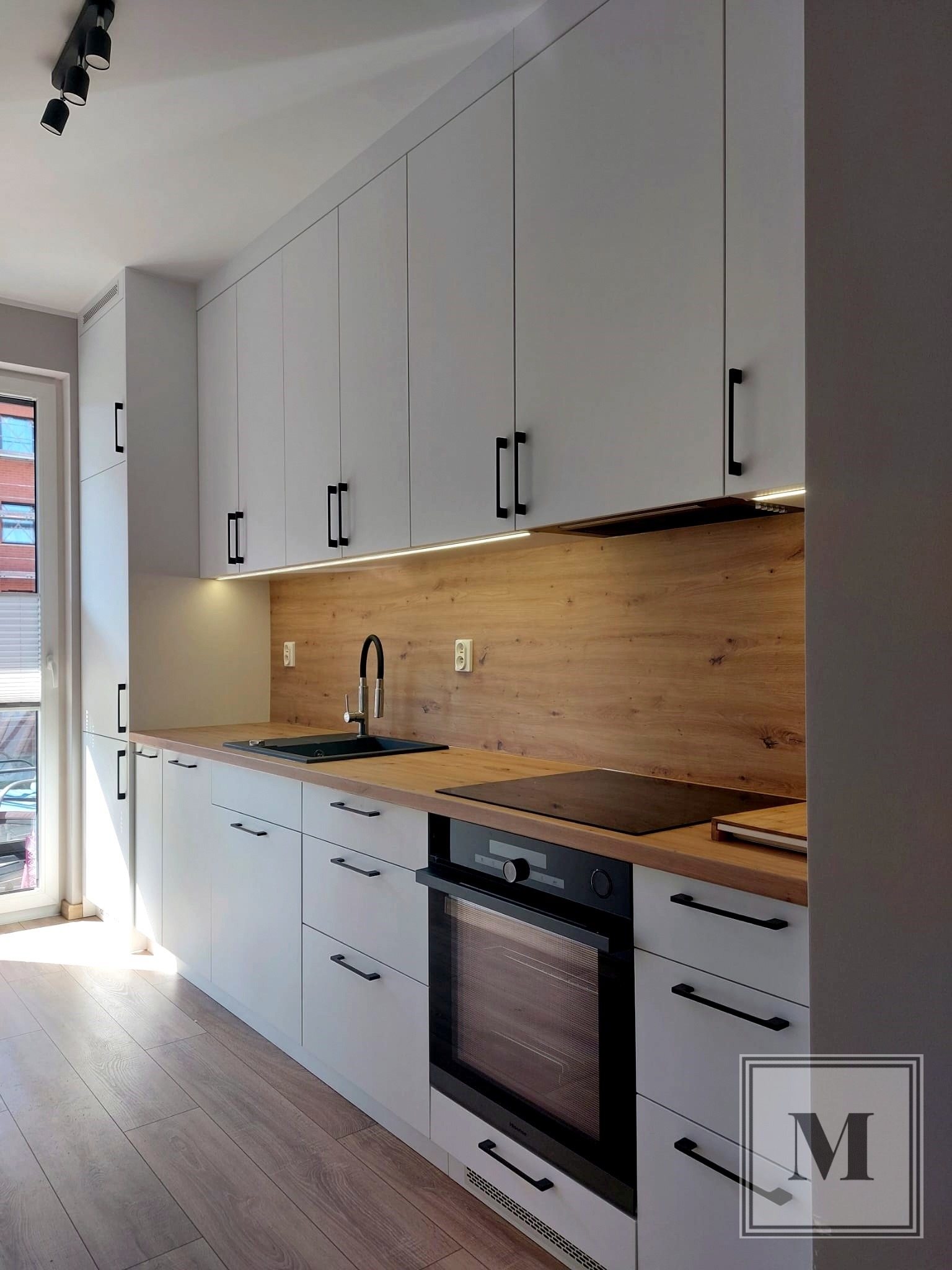 Nowoczesna kuchnia z białymi szafkami, drewnianym blatem i panelem ściennym, czarną armaturą i oświetleniem LED podszafkowym.