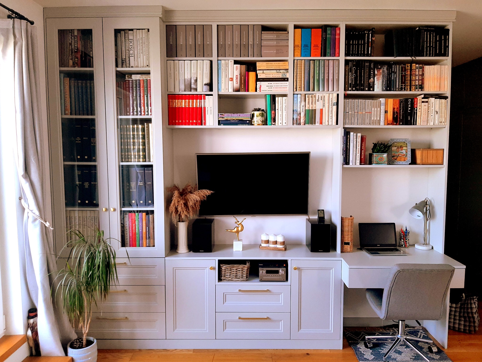 Szafa wnękowa z szarymi frontami, wbudowana w ścianę, z otwartymi półkami na książki i zamkniętymi szafkami, z biurkiem po prawej stronie, telewizorem pośrodku i dekoracjami, całość oświetlona...