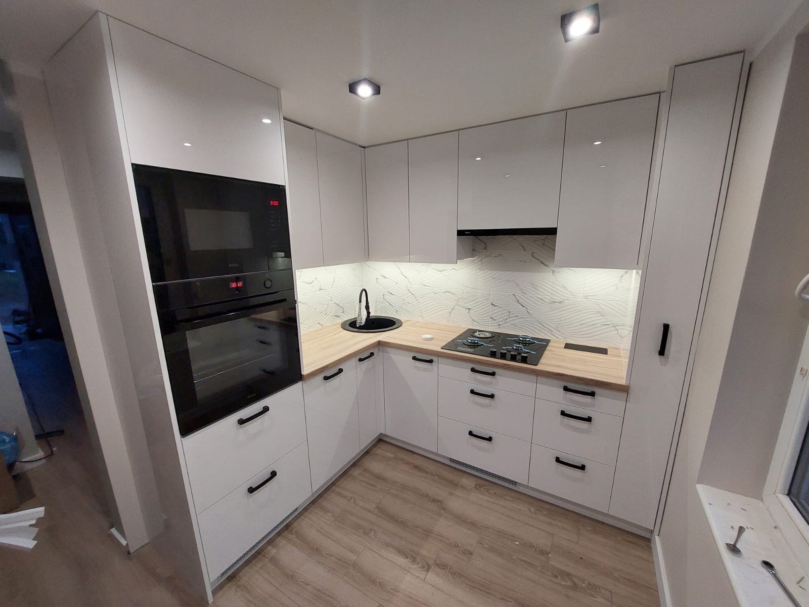 Nowoczesna, jasna kuchnia z białymi szafkami z połyskiem, drewnianym blatem i czarnymi uchwytami, wyposażona w płytę grzewczą i piekarnik w zabudowie.