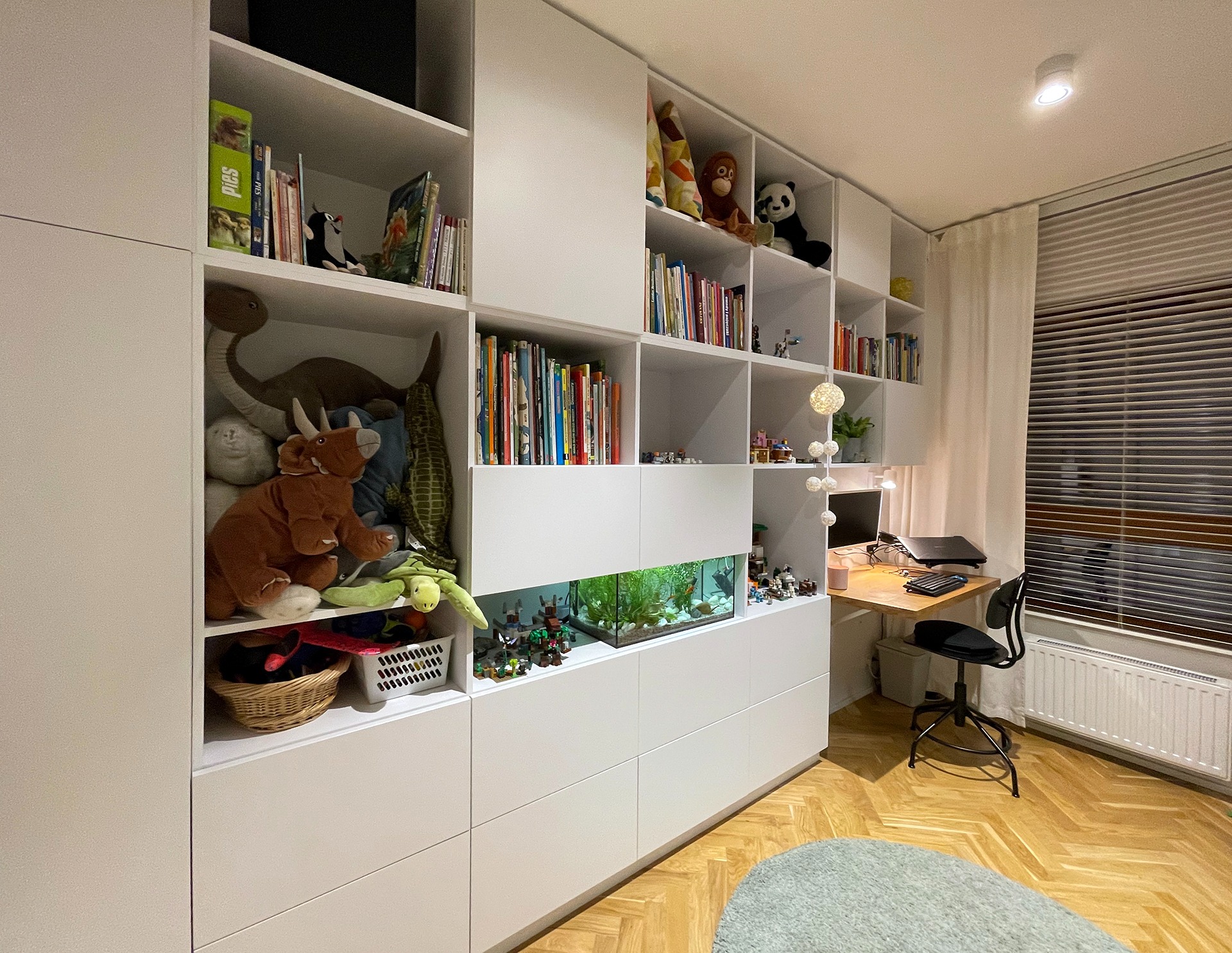 Biała zabudowa meblowa w pokoju dziecięcym z otwartymi półkami, na których znajdują się książki, zabawki i akwarium z rybkami.