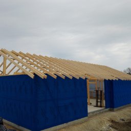 BUDODACH - Budowa domu w stanie surowym otwartym: drewniana konstrukcja dachu, ściany pokryte niebieską membraną, betonowa podmurówka. Widok z zewnątrz, pochmurne niebo.