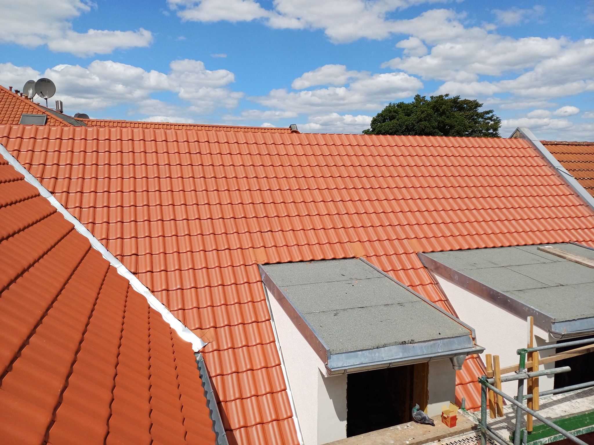 Czerwona dachówka ceramiczna na dachu z lukarnami, widok z góry w słoneczny dzień. Na dachu widoczne elementy wykończeniowe i fragment rusztowania.