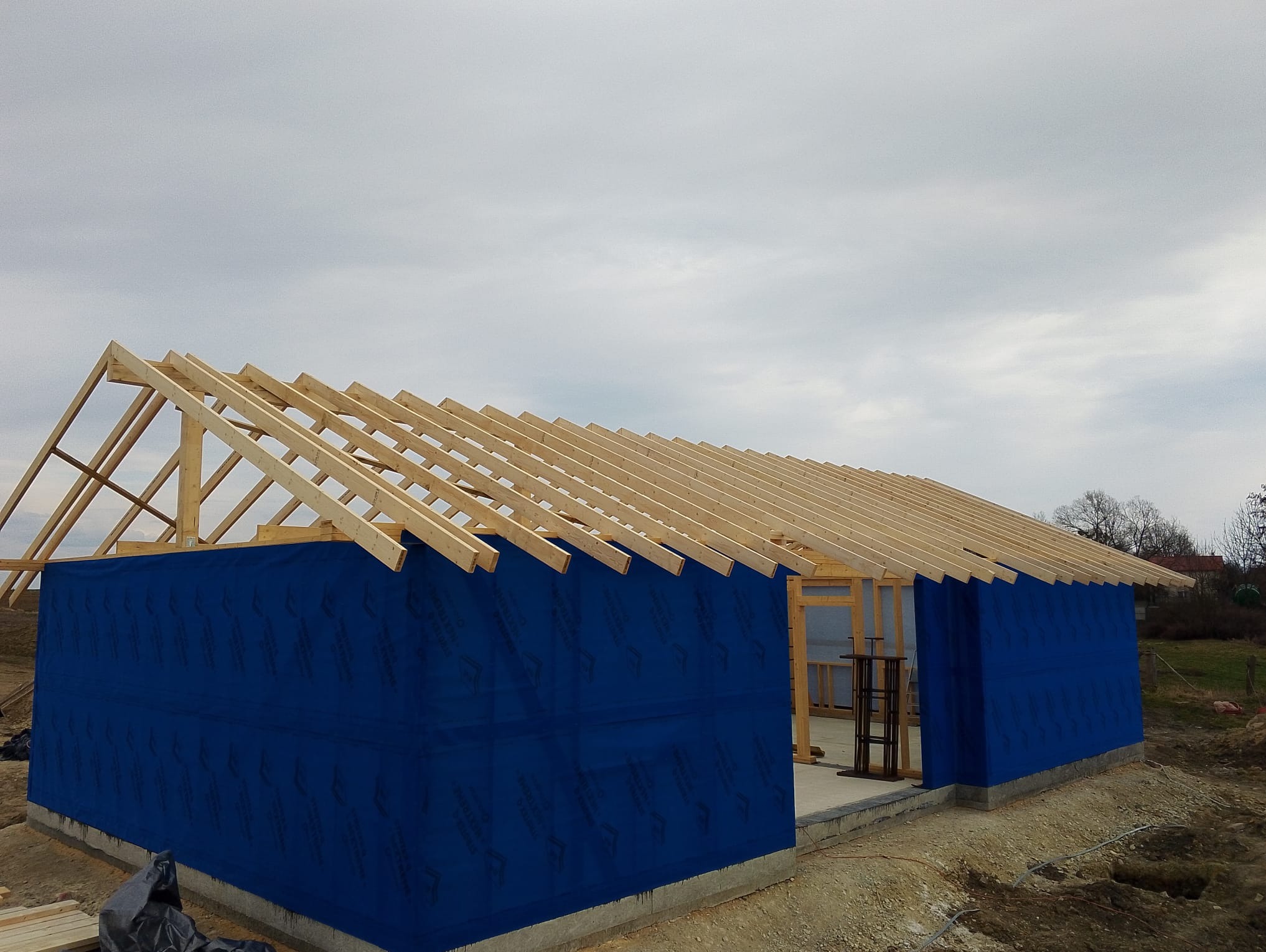 Budowa domu w stanie surowym otwartym: drewniana konstrukcja dachu, ściany pokryte niebieską membraną, betonowa podmurówka. Widok z zewnątrz, pochmurne niebo.