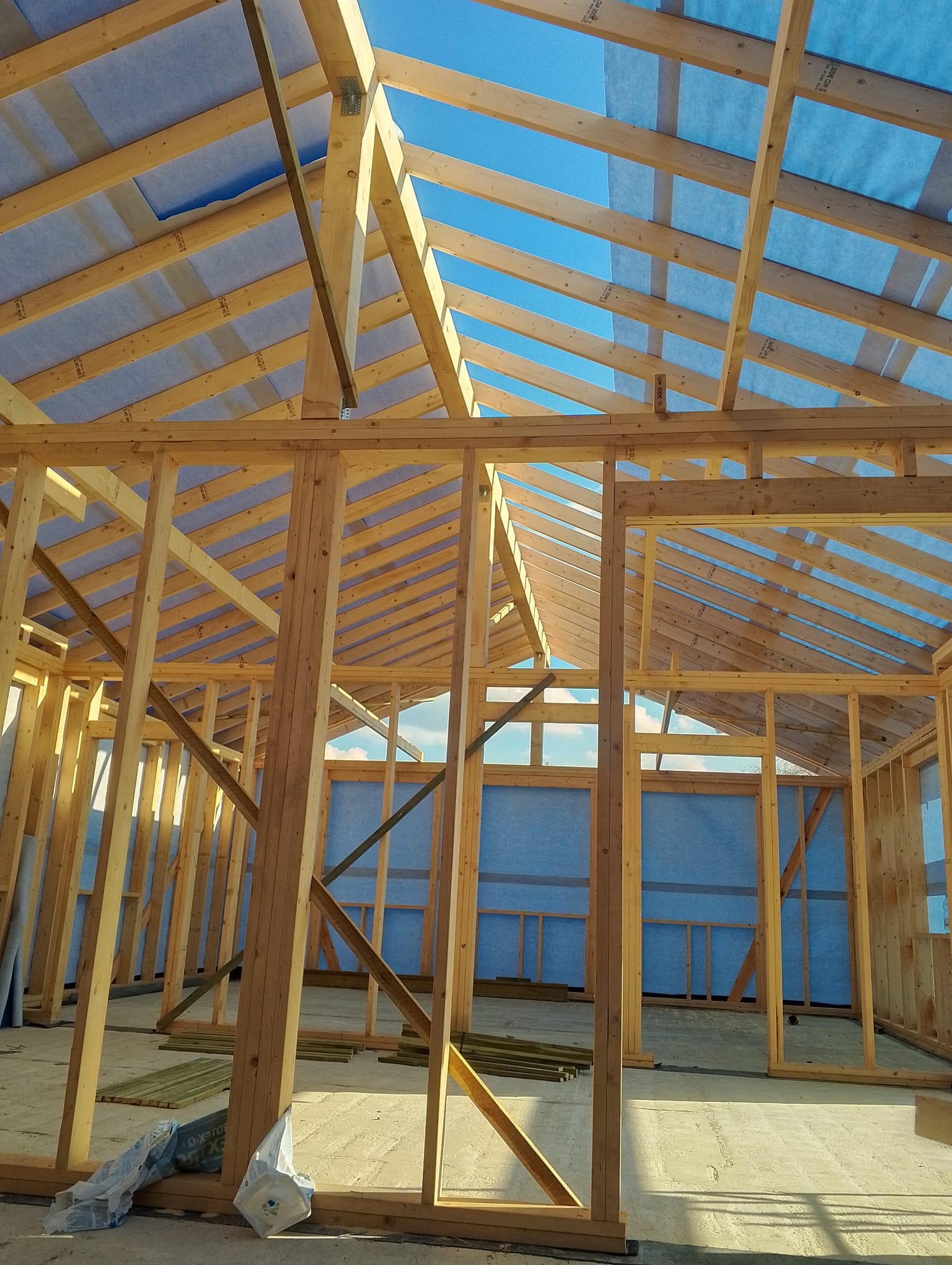 Wnętrze domu w trakcie budowy z drewnianą konstrukcją szkieletową i niebieską membraną dachową, widok z dołu na niebo przez otwory w dachu.