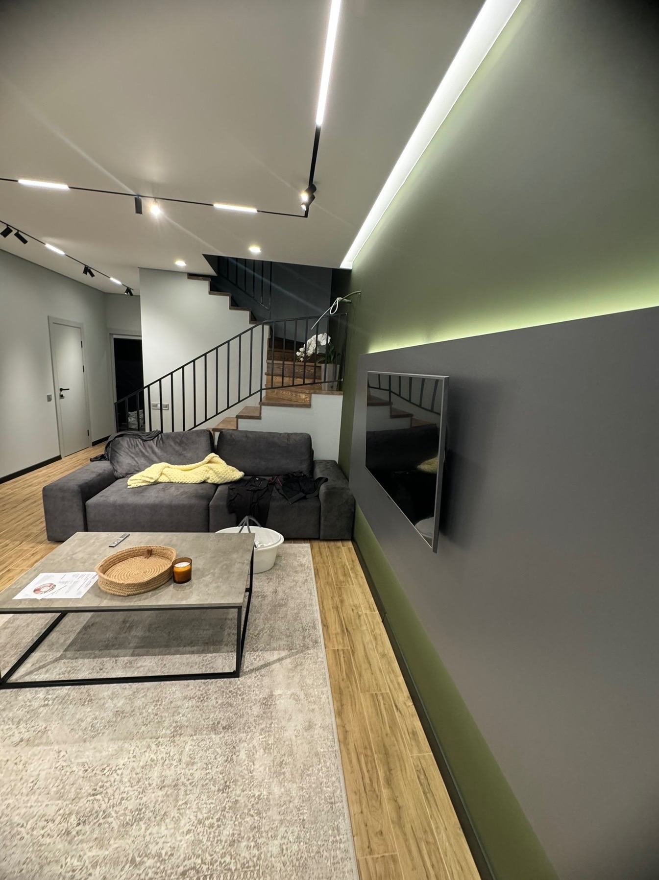 Nowoczesny salon z szarą sofą, drewnianymi schodami i podświetlaną ścianą. Minimalistyczny design z akcentami światła i cienia. Widoczny telewizor na ścianie.