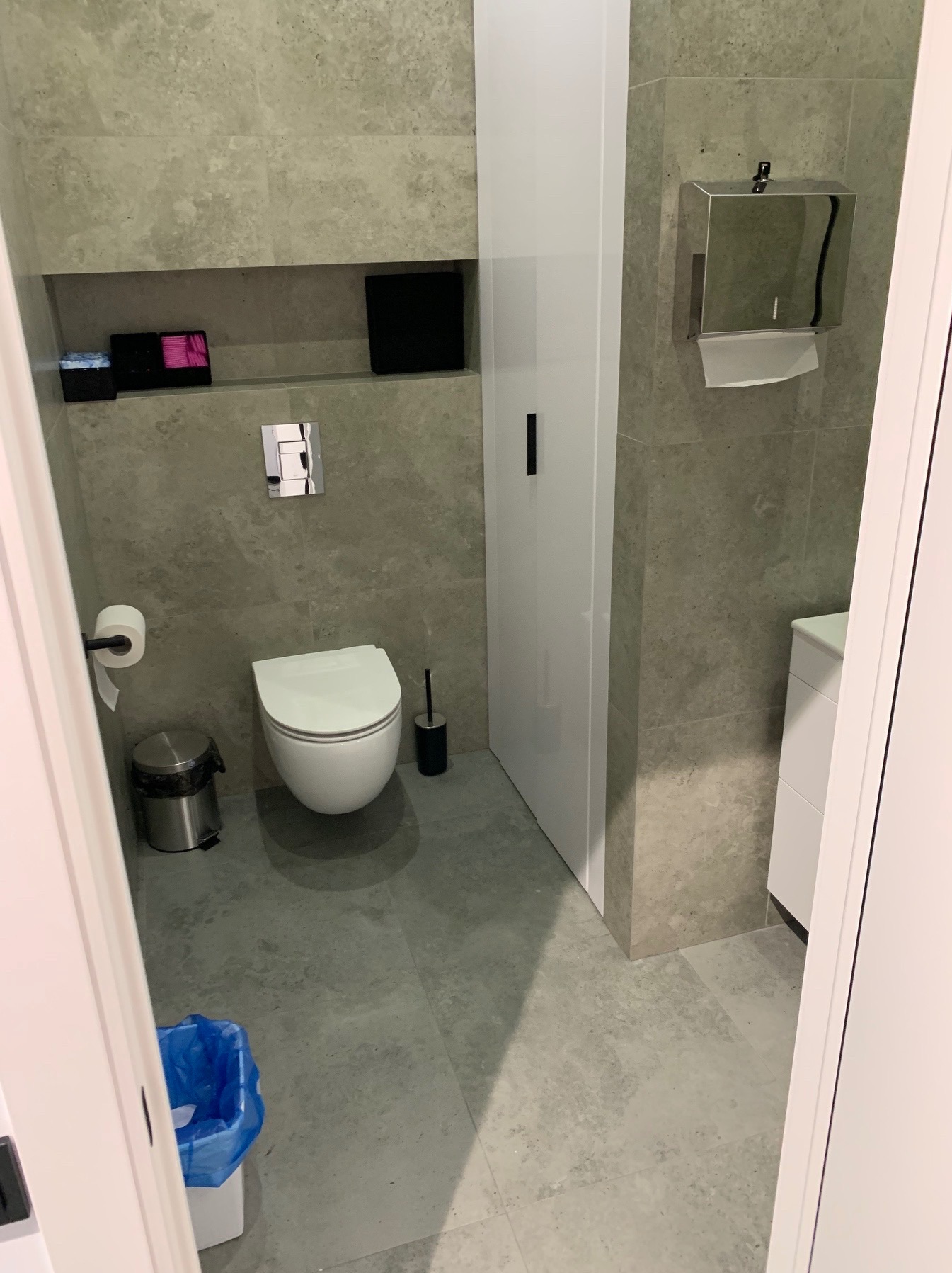 Nowoczesna łazienka z szarymi płytkami imitującymi kamień, podwieszana toaleta, biała szafka w zabudowie i metalowy podajnik na ręczniki. Minimalistyczny design.