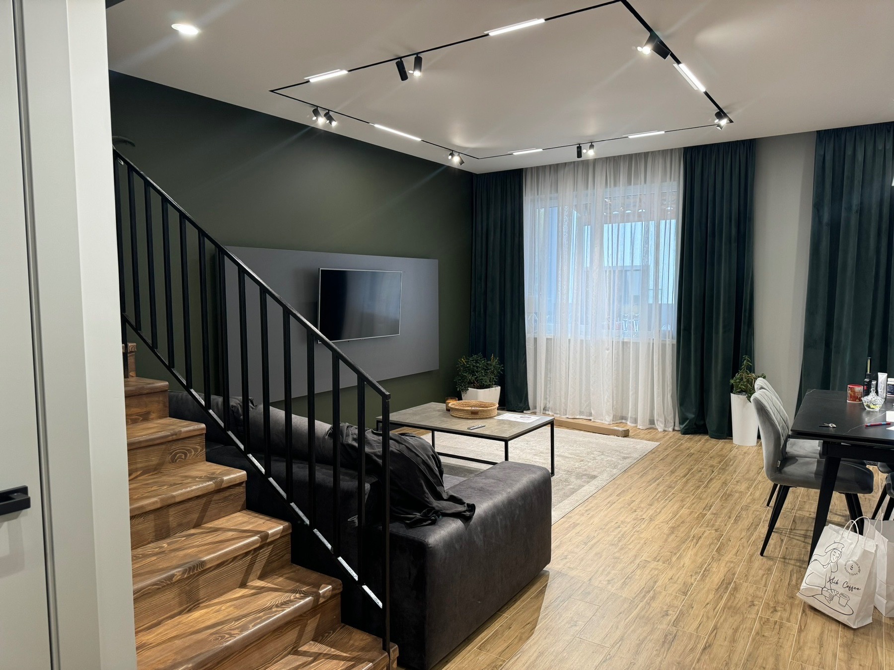 Nowoczesne wnętrze: salon z drewnianymi schodami, ciemnozieloną ścianą, szarą sofą i oświetleniem szynowym. Widoczne okno z zasłonami i stół z krzesłami.