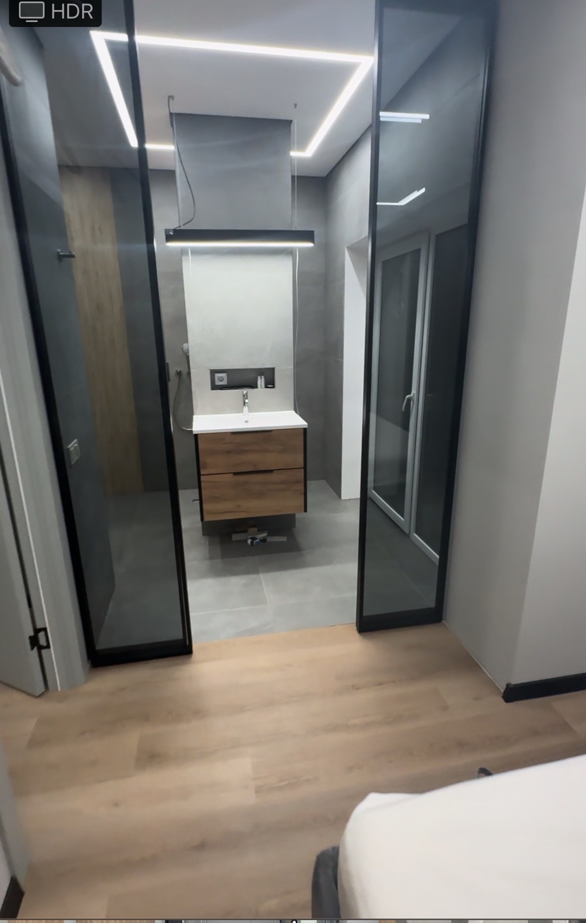 Nowoczesna łazienka z drewnianą szafką pod umywalką, szarą glazurą i oświetleniem LED, widziana przez szklane drzwi.