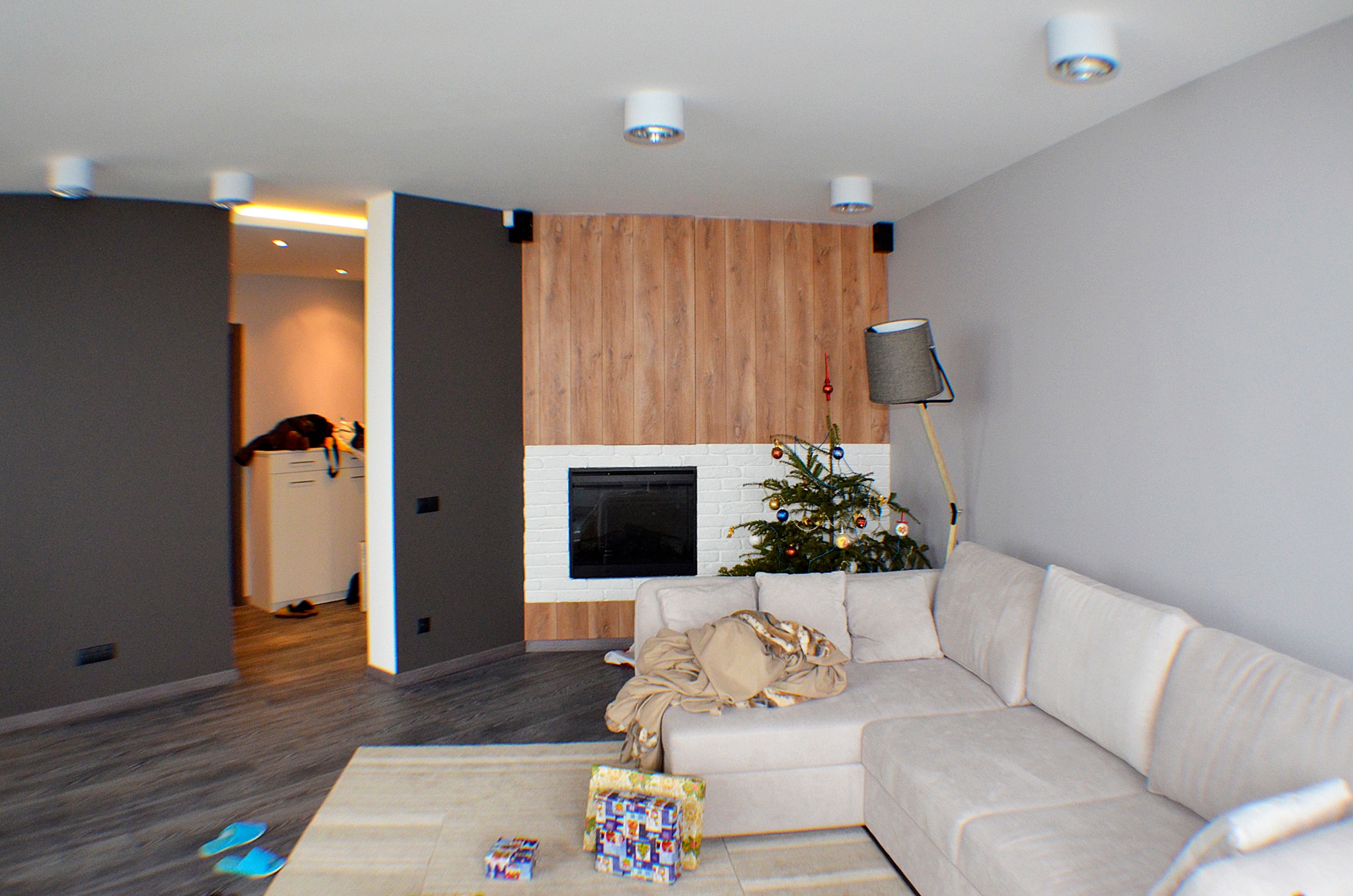Jasne wnętrze salonu z kominkiem obłożonym białą cegłą i drewnianą ścianą, narożna sofa z narzutą, choinka i prezenty, widok na sąsiednie pomieszczenie z szarymi ścianami.