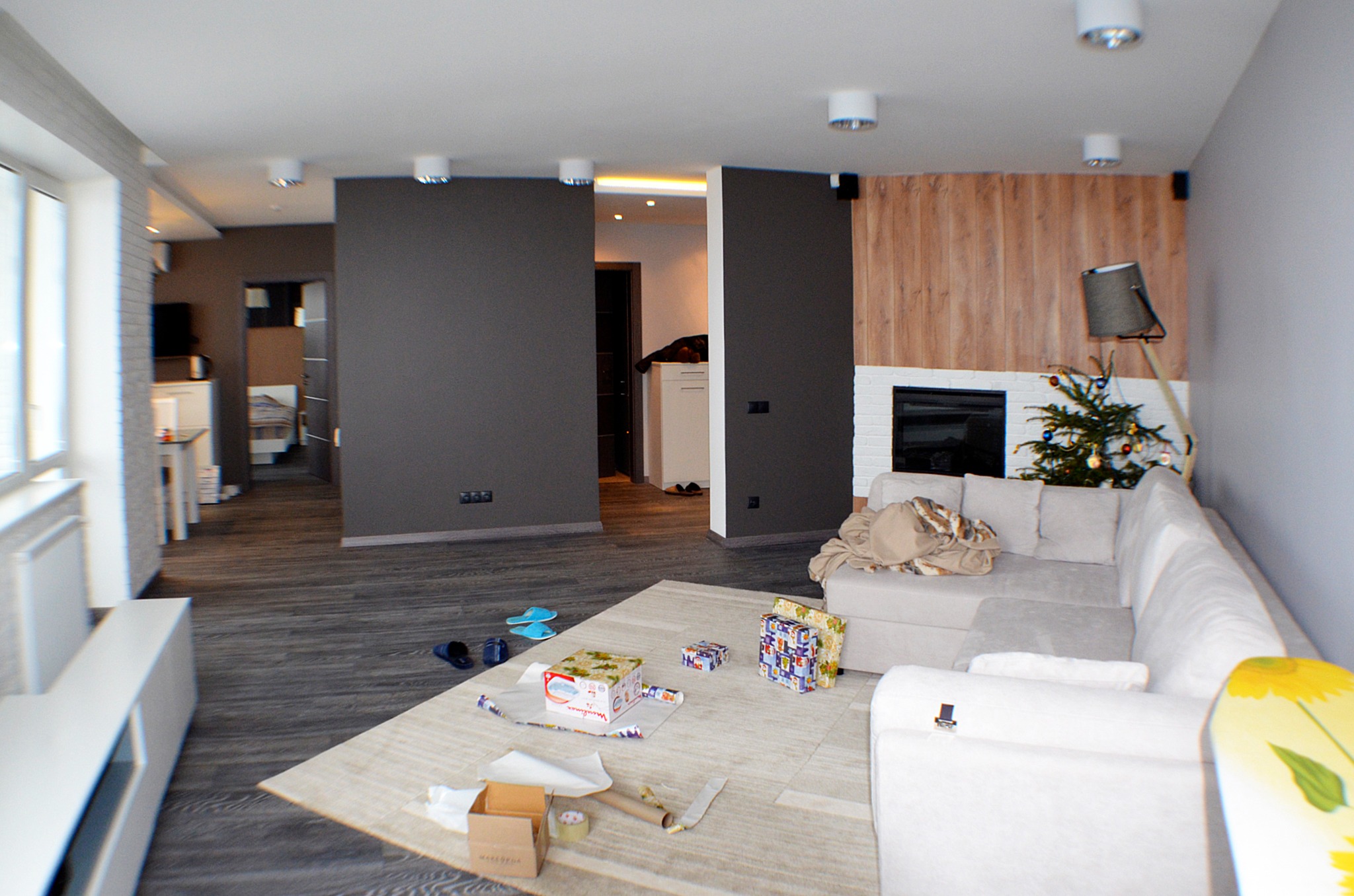 Przestronne wnętrze salonu z drewnianą podłogą i elementami dekoracyjnymi: choinka, prezenty na dywanie, przesuwne drzwi w kolorze grafitowym, biała sofa narożna i telewizor.