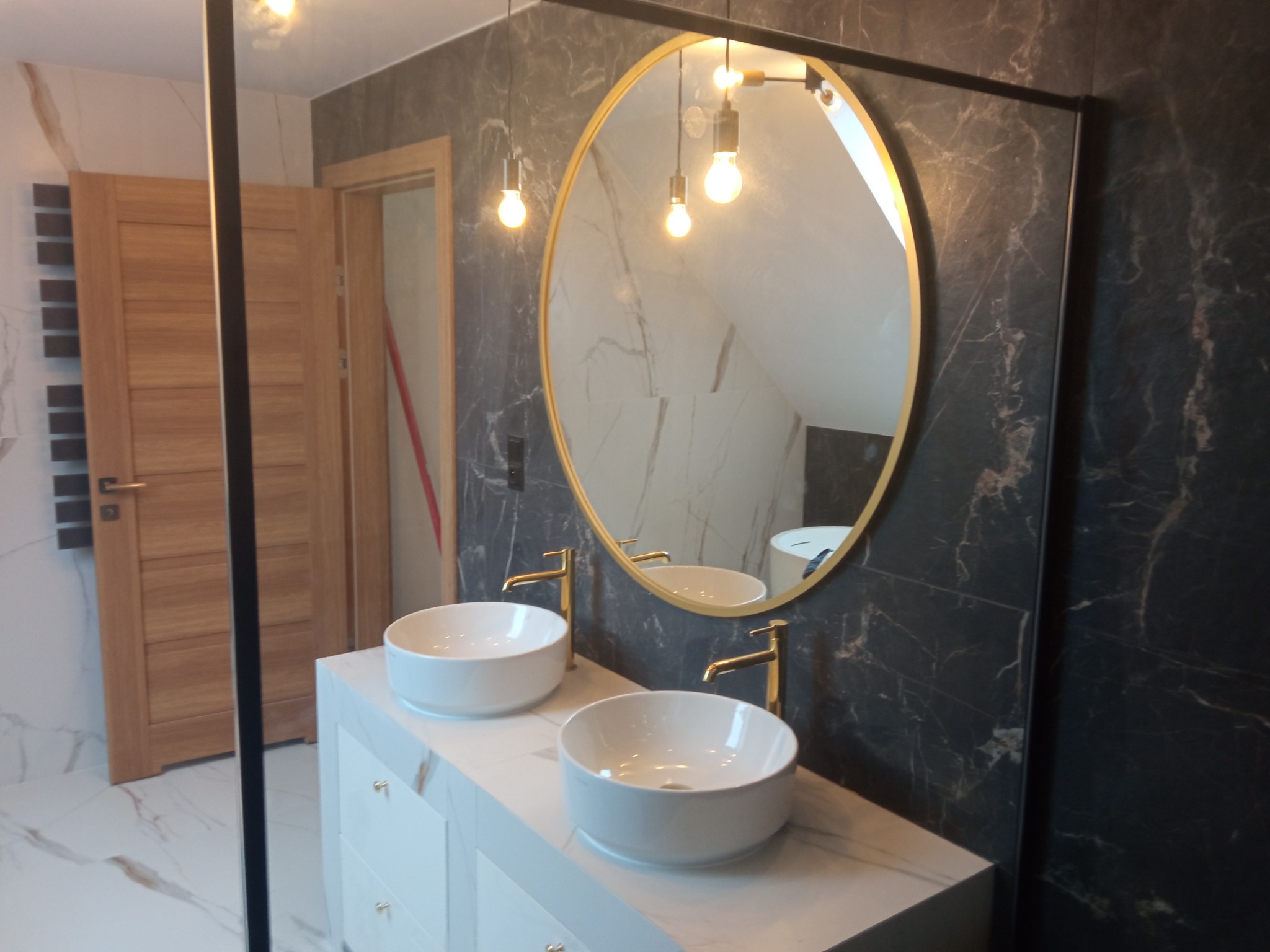 Nowoczesna łazienka z dwoma okrągłymi umywalkami nablatowymi z złotymi bateriami, marmurowe ściany i podłoga, owalne lustro z złotą ramą i wiszące lampy z żarówkami Edisona.