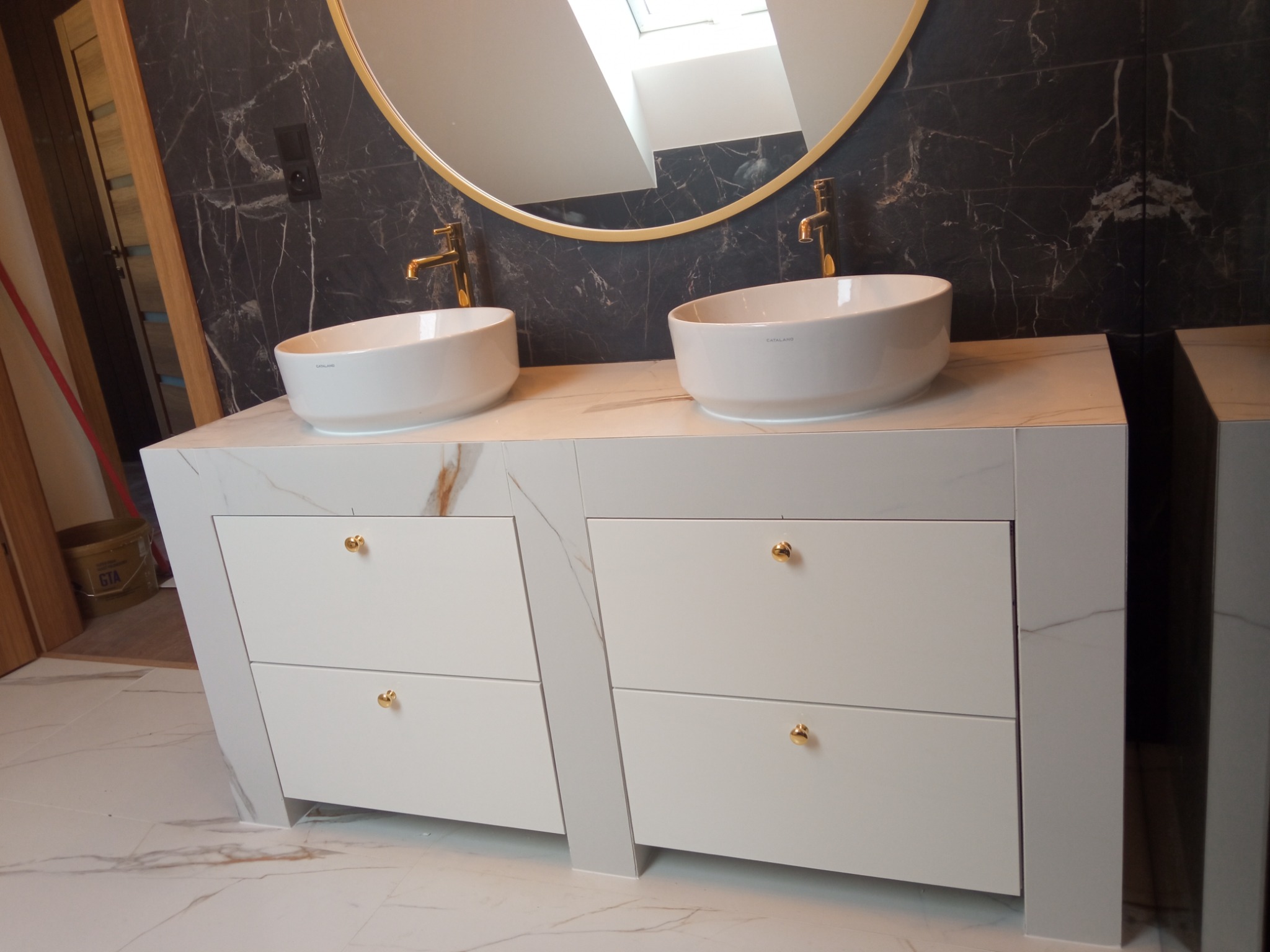 Nowoczesna łazienka z dwoma białymi umywalkami nablatowymi, złotymi bateriami i okrągłym lustrem w złotej ramie na tle ściany wyłożonej czarnymi płytkami imitującymi marmur.