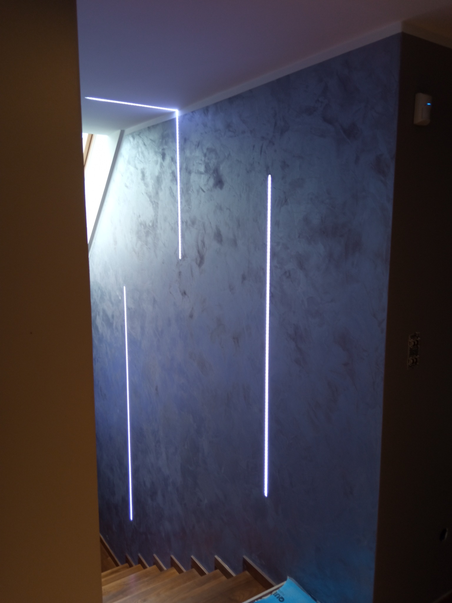 Ściana w korytarzu z dekoracyjną, szarą strukturą i pionowymi, wpuszczonymi liniami LED, oświetlającymi drewniane schody.