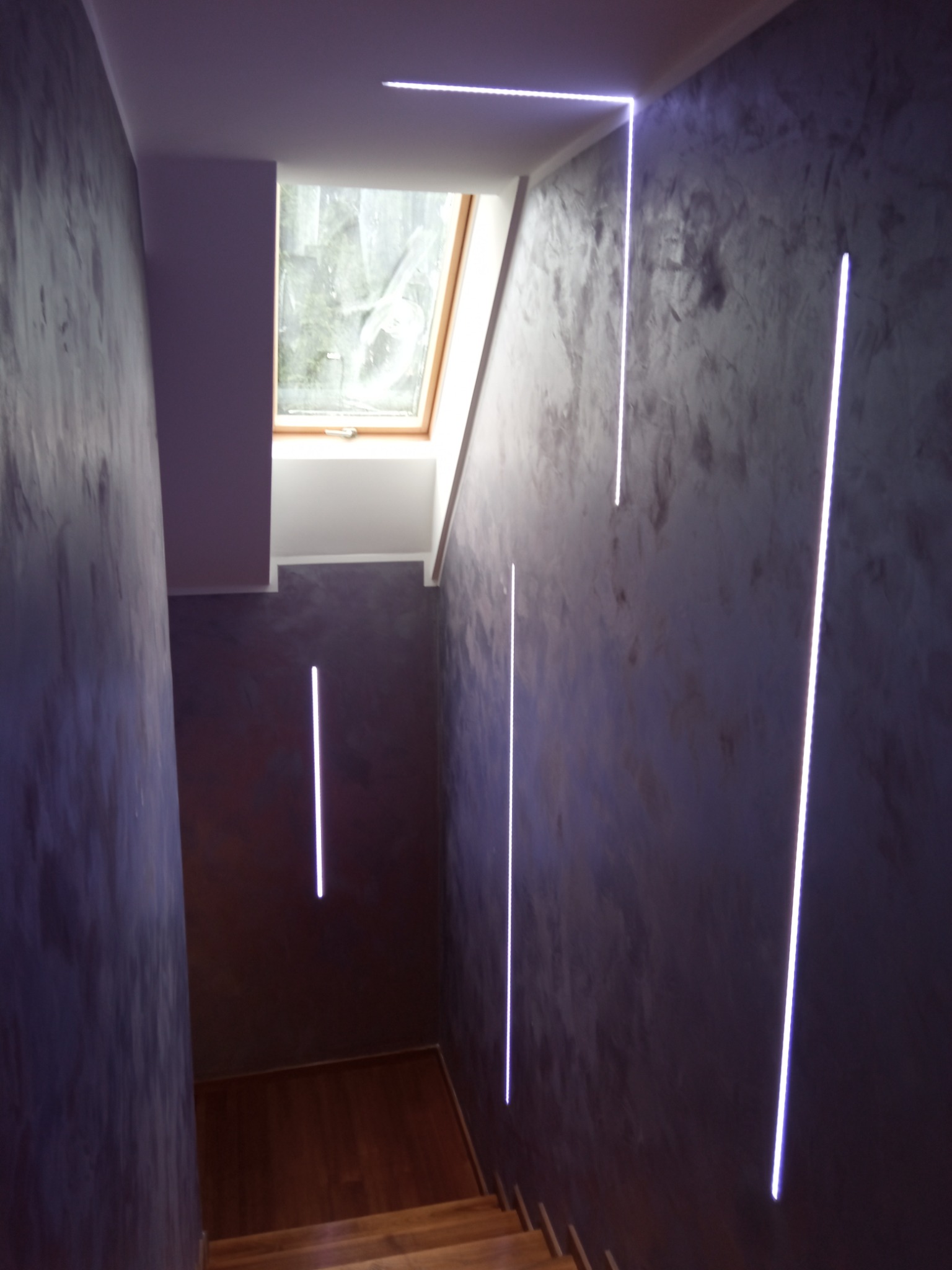 Klatka schodowa z dekoracyjnymi pionowymi paskami LED na ścianach o fakturze betonu i drewnianymi schodami prowadzącymi na poddasze z oknem dachowym.