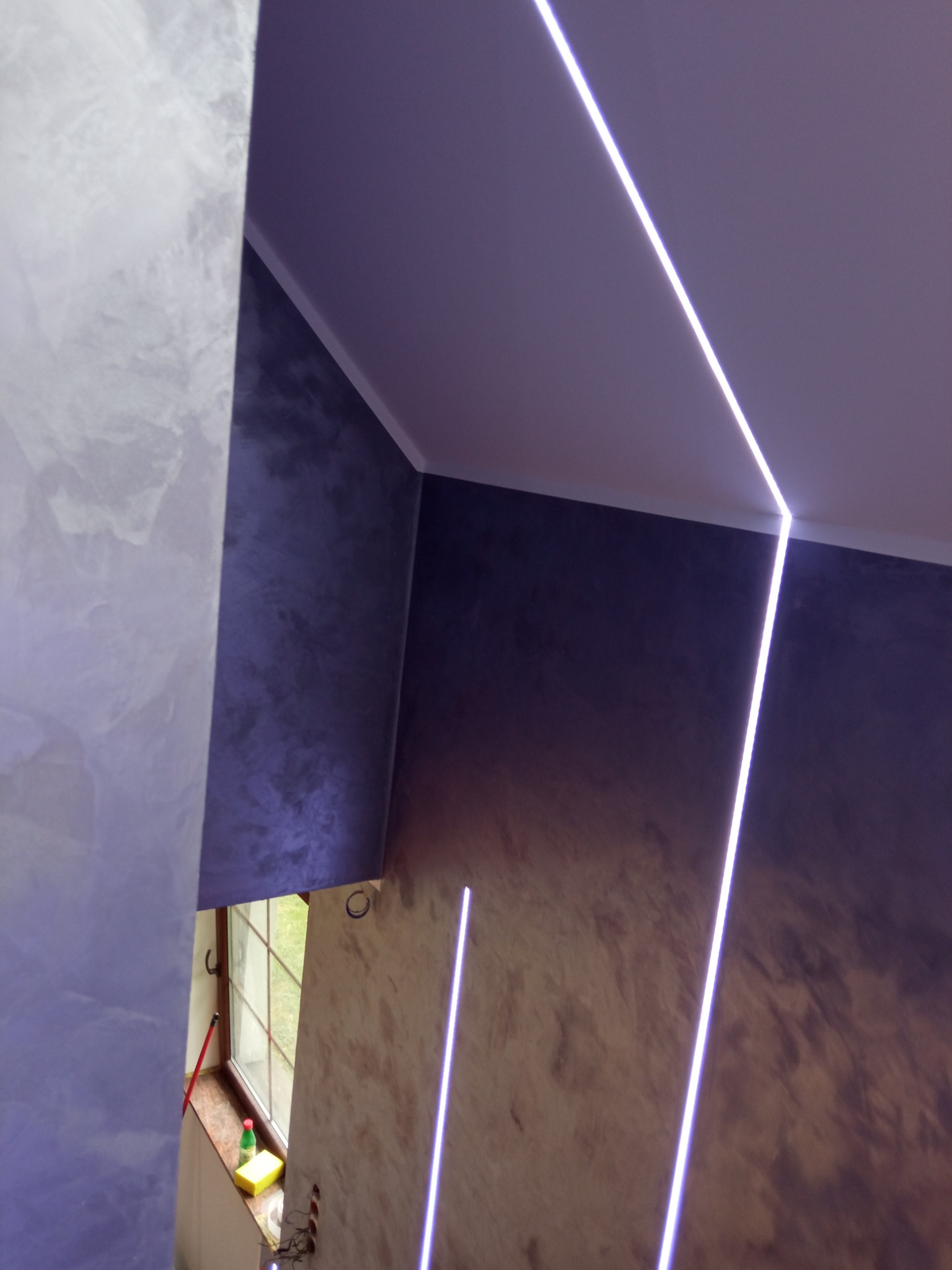 Ściany w pokoju wykończone w odcieniach szarości i fioletu z dekoracyjnym oświetleniem LED wzdłuż sufitu i pionowo w rogu, widoczne okno z parapetem i akcesoriami do czyszczenia.