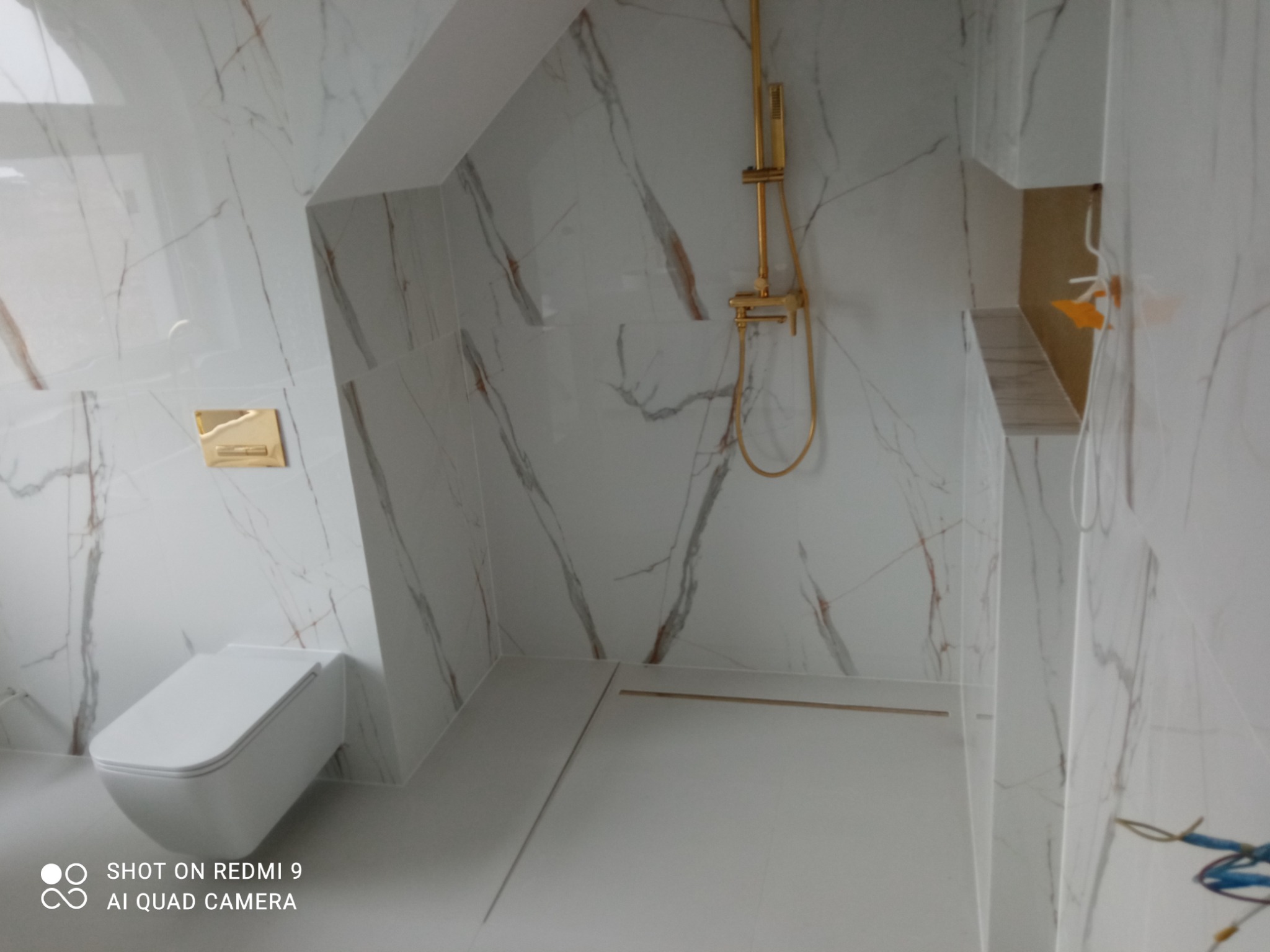 Nowoczesna łazienka w bieli z marmurowym wzorem na ścianach, wyposażona w złotą armaturę prysznicową i podwieszaną toaletę.
