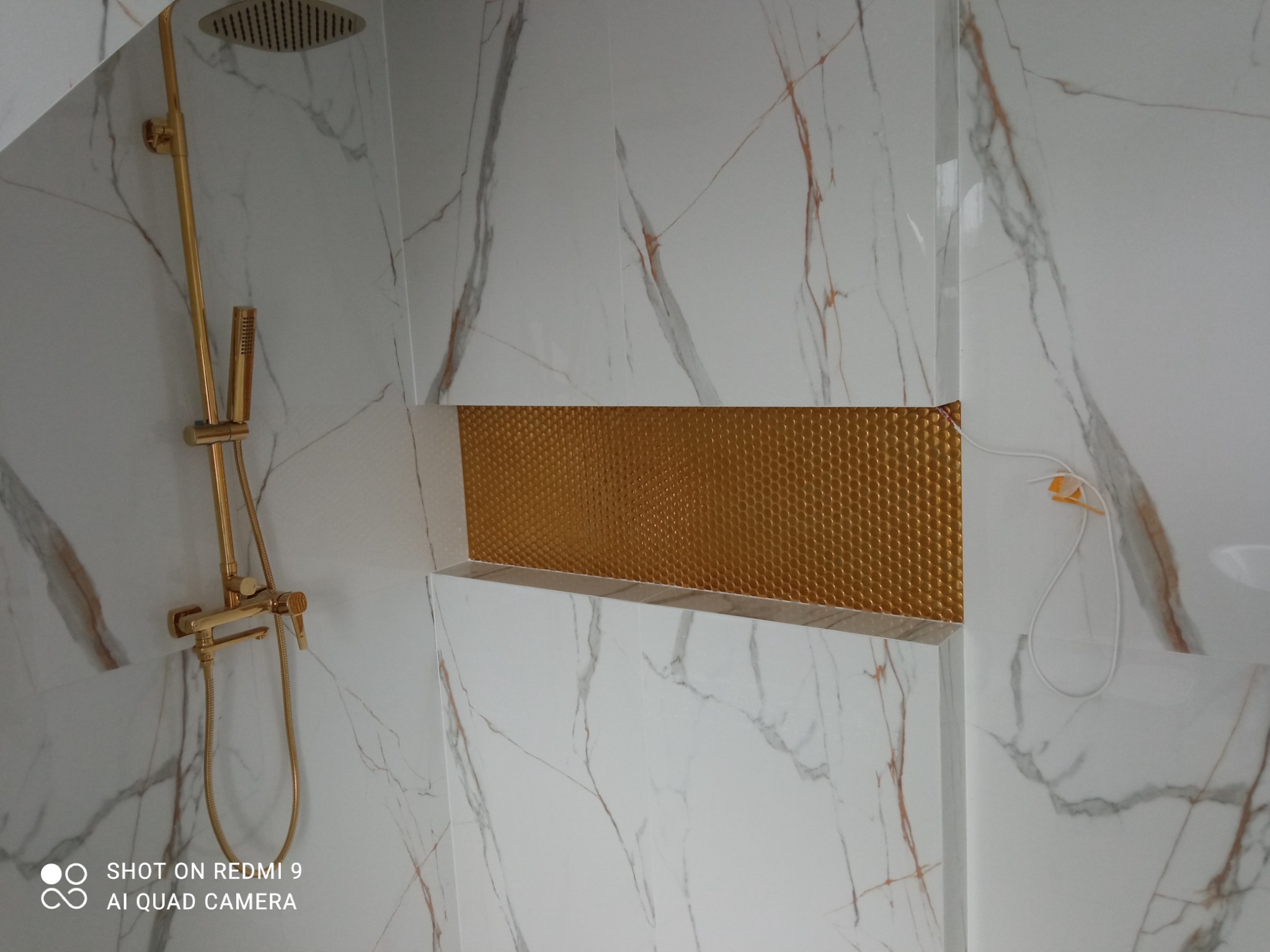 Wykończona łazienka z białymi płytkami imitującymi marmur, złotą armaturą prysznicową i wnęką wyłożoną złotą mozaiką o strukturze plastra miodu.