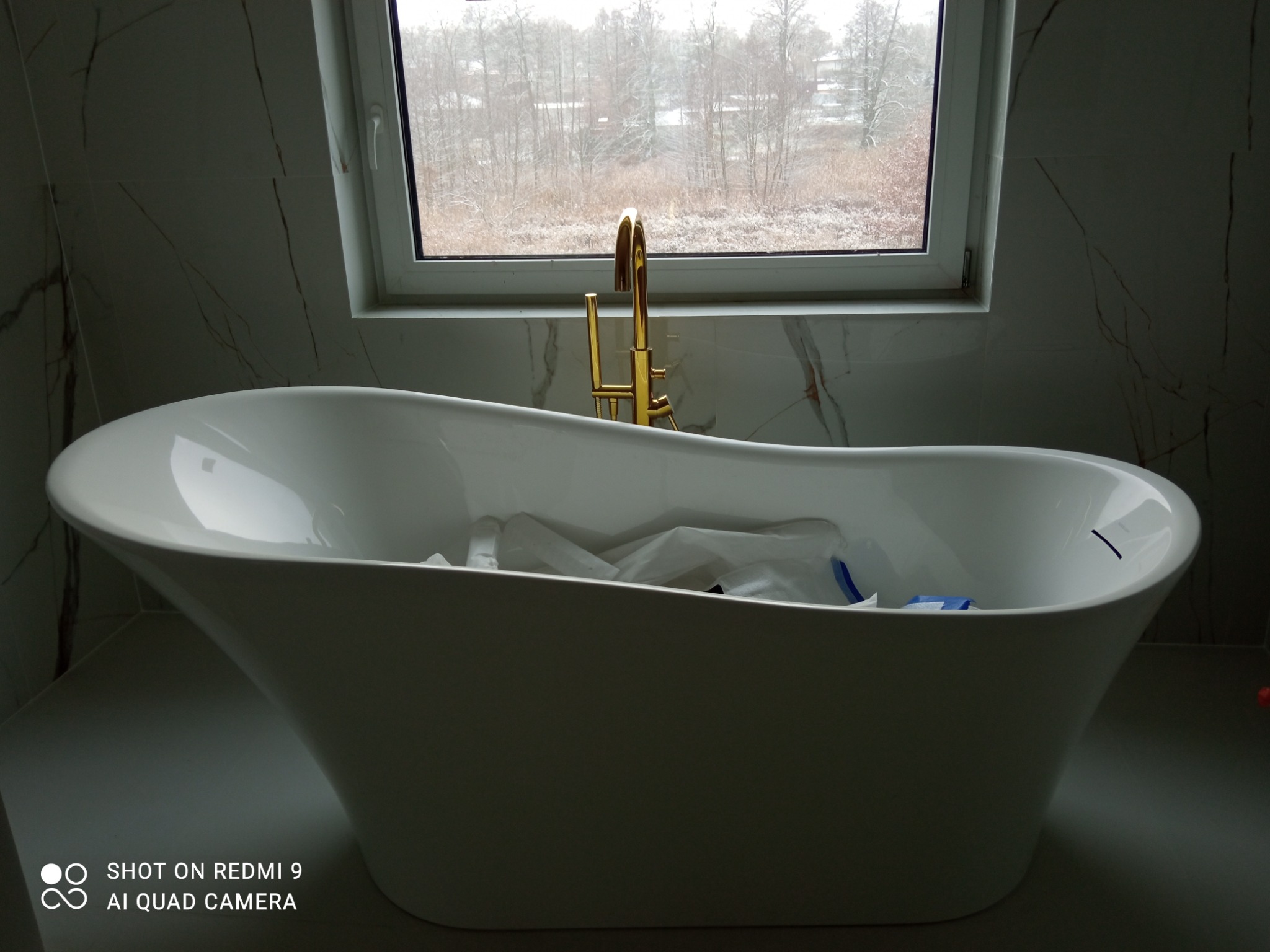 Wolnostojąca wanna z złotą armaturą pod oknem z widokiem na zimowy krajobraz, białe marmurowe płytki na ścianie.