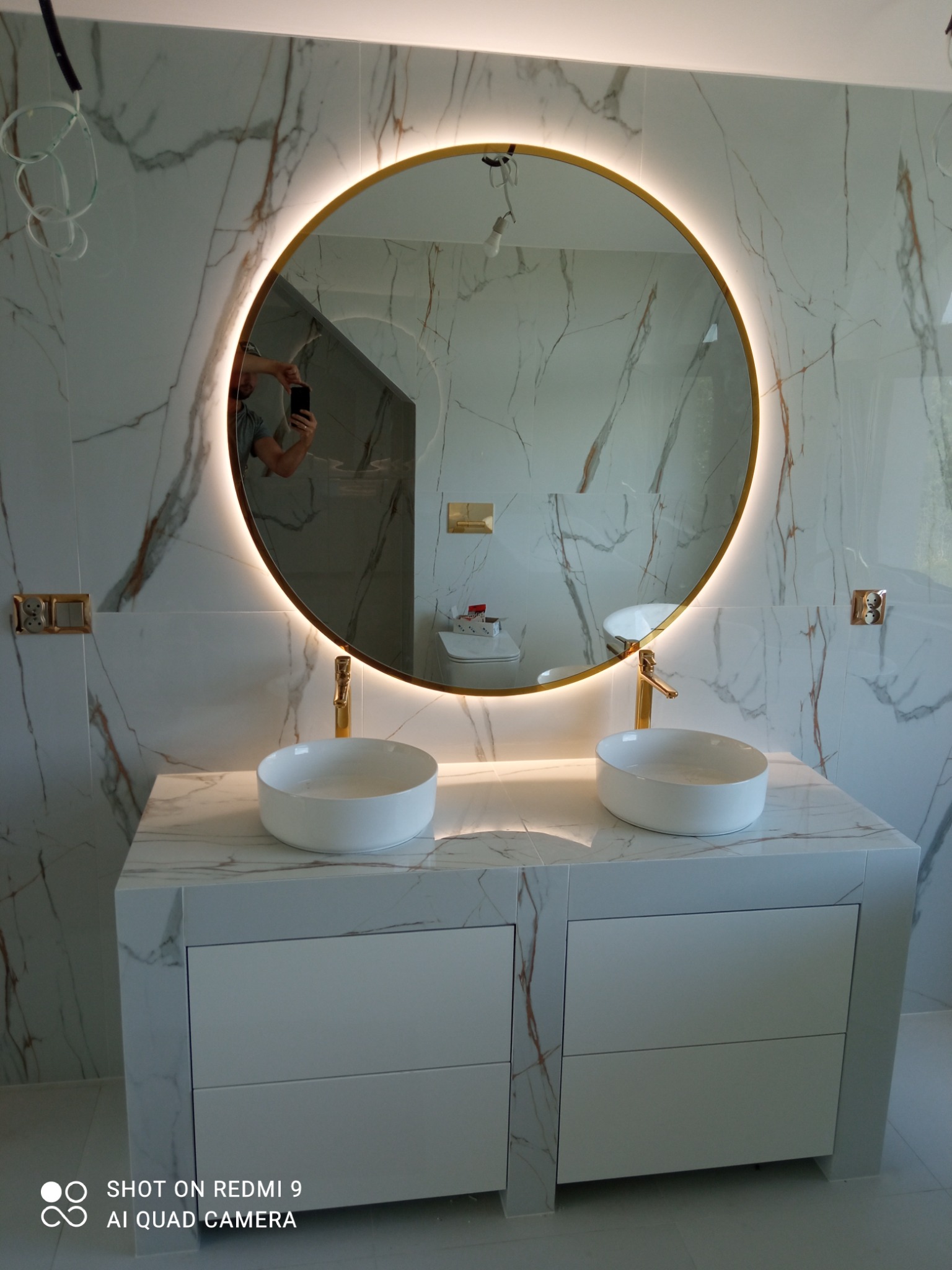 Łazienka z dwoma umywalkami nablatowymi, okrągłe lustro z podświetleniem LED, marmurowe płytki na ścianach i blacie, białe szafki pod umywalkami.