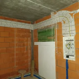 Surowy stan: Kable elektryczne ułożone w korytkach na ścianach z cegły dziurawki, z widoczną skrzynką rozdzielczą i fragmentem instalacji w niebieskiej rurze karbowanej.