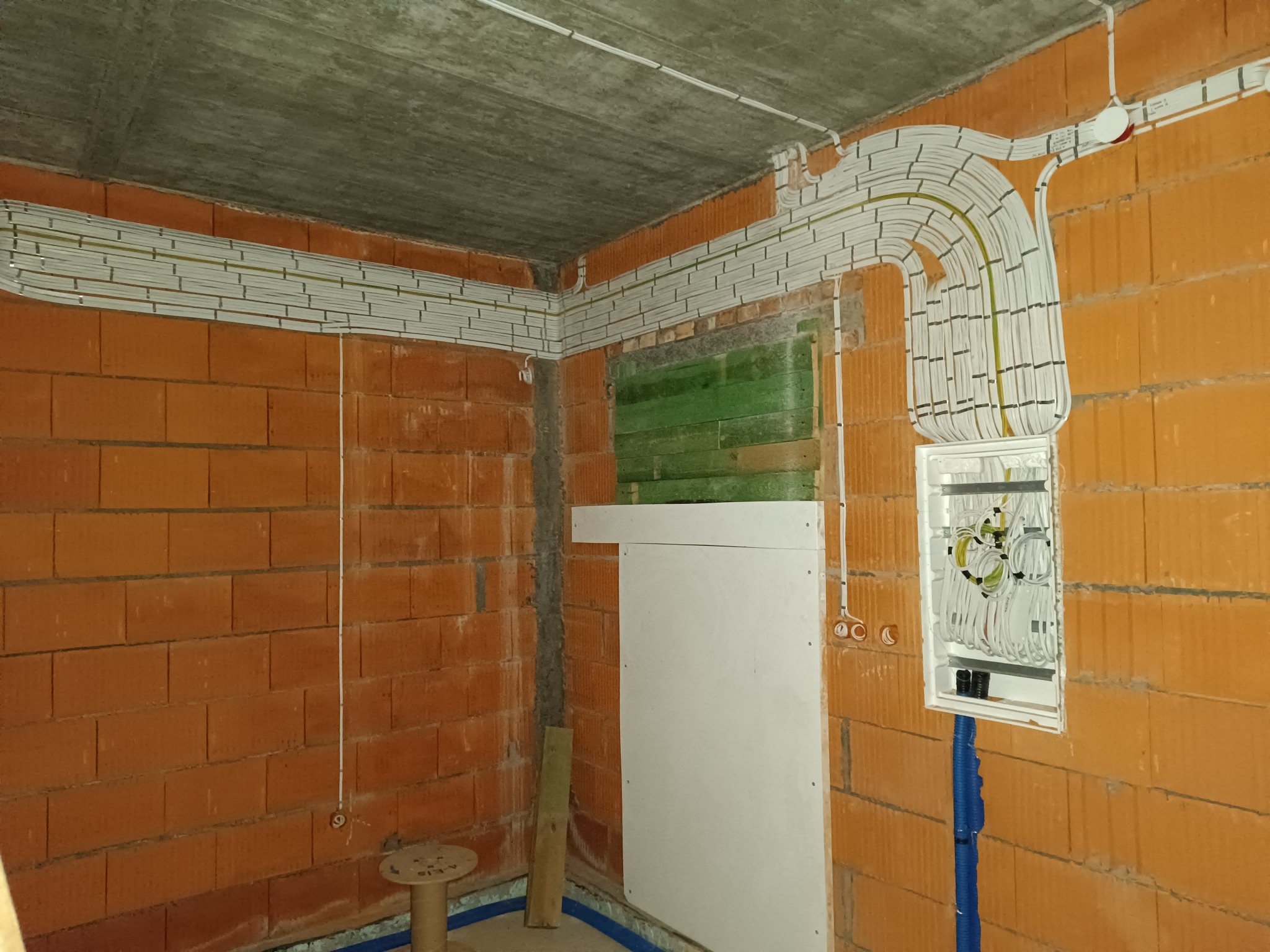 Surowy stan: Kable elektryczne ułożone w korytkach na ścianach z cegły dziurawki, z widoczną skrzynką rozdzielczą i fragmentem instalacji w niebieskiej rurze karbowanej.