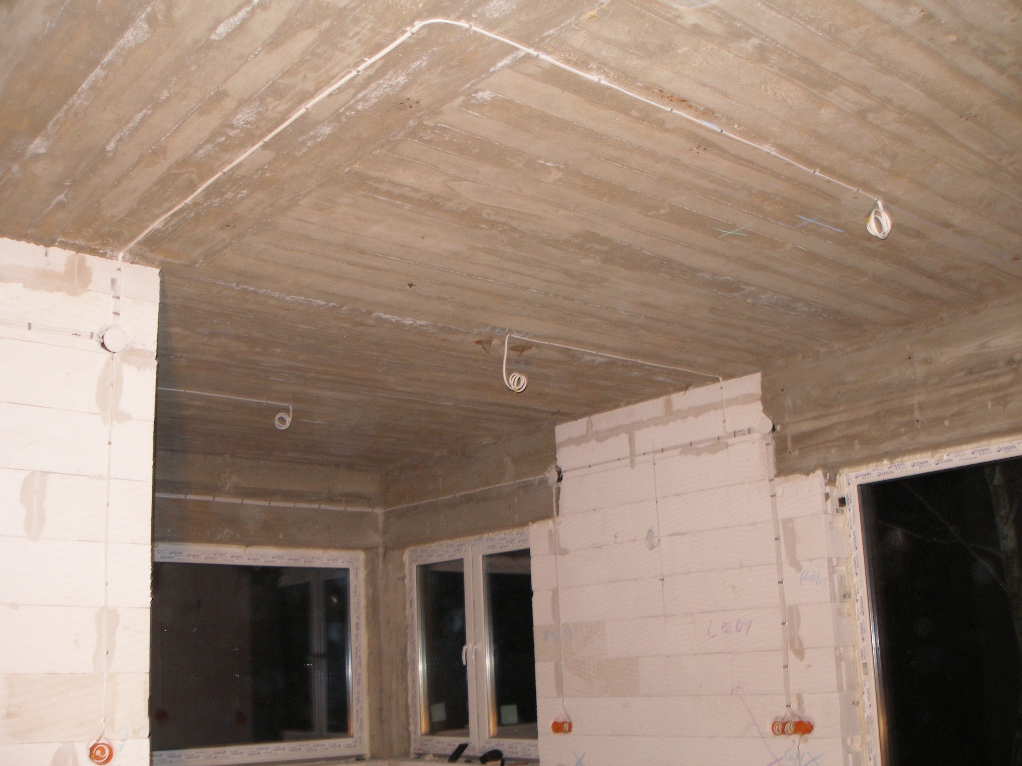 Surowy betonowy sufit z widocznymi przewodami elektrycznymi i punktami oświetleniowymi w trakcie instalacji, ściany z bloków z betonu komórkowego przed wykończeniem, okna w stanie deweloperskim.