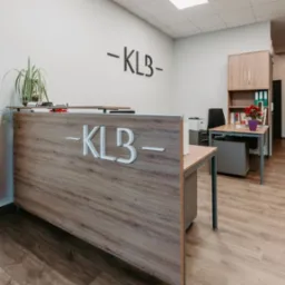 Jasne wnętrze biura z recepcją z drewnianym frontem, na kt&oacute;rej widnieje białe logo firmy -KLB-, takie samo logo w czarnym kolorze na ścianie, biurko z szafką i regał z segregatorami w tle.