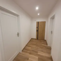 Wykończony korytarz z panelami podłogowymi imitującymi drewno, białe ściany i trzy pary drzwi: dwie białe wewnętrzne i jedna drewnopodobna wejściowa z czarnymi akcentami.