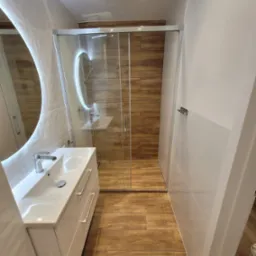 Nowoczesna łazienka z kabiną prysznicową typu walk-in, umywalką z szafką w kolorze białym i okrągłym lustrem z podświetleniem LED.