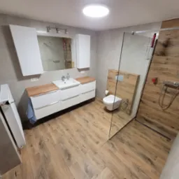 Nowoczesna łazienka w Glisno z kabiną prysznicową typu walk-in, drewnopodobnymi płytkami na podłodze i ścianie, białymi szafkami pod umywalką z drewnianym blatem oraz wiszącą toaletą.