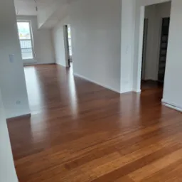 Błyszcząca, nowa podłoga z ciemnego drewna, prawdopodobnie parkiet, w jasnym, jeszcze niewykończonym pokoju z białymi ścianami i wystającymi kablami od lamp.