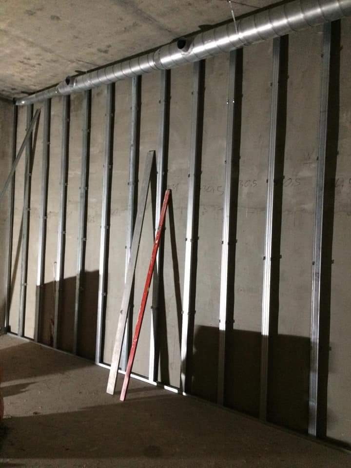 Konstrukcja ścianki działowej z profili stalowych i płyt gipsowo-kartonowych w surowym stanie, widoczne przewody wentylacyjne pod betonowym sufitem, poziomica oparta o konstrukcję.