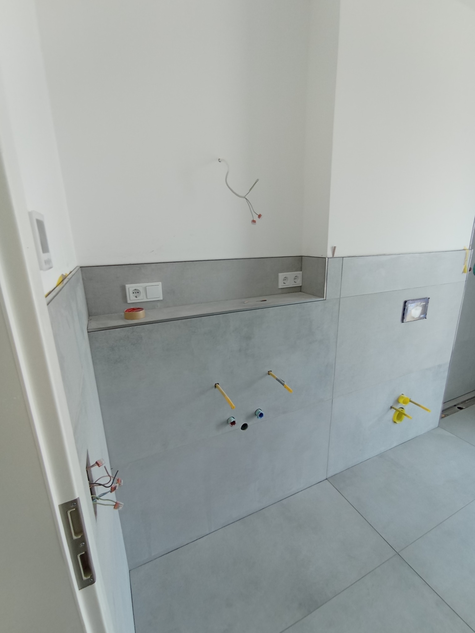 Szare płytki wielkoformatowe ułożone na ścianach i podłodze w trakcie wykańczania łazienki, widoczne wystające rury i kable, puszki elektryczne oraz taśma malarska.