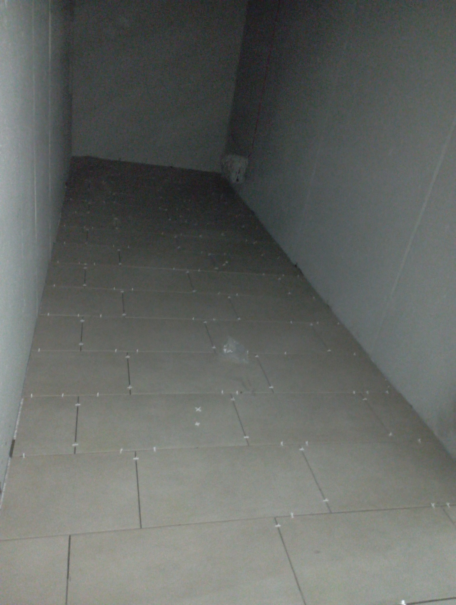 Układanie jasnoszarych płytek na podłodze, widoczne krzyżyki dystansowe, surowy stan, słabe oświetlenie.