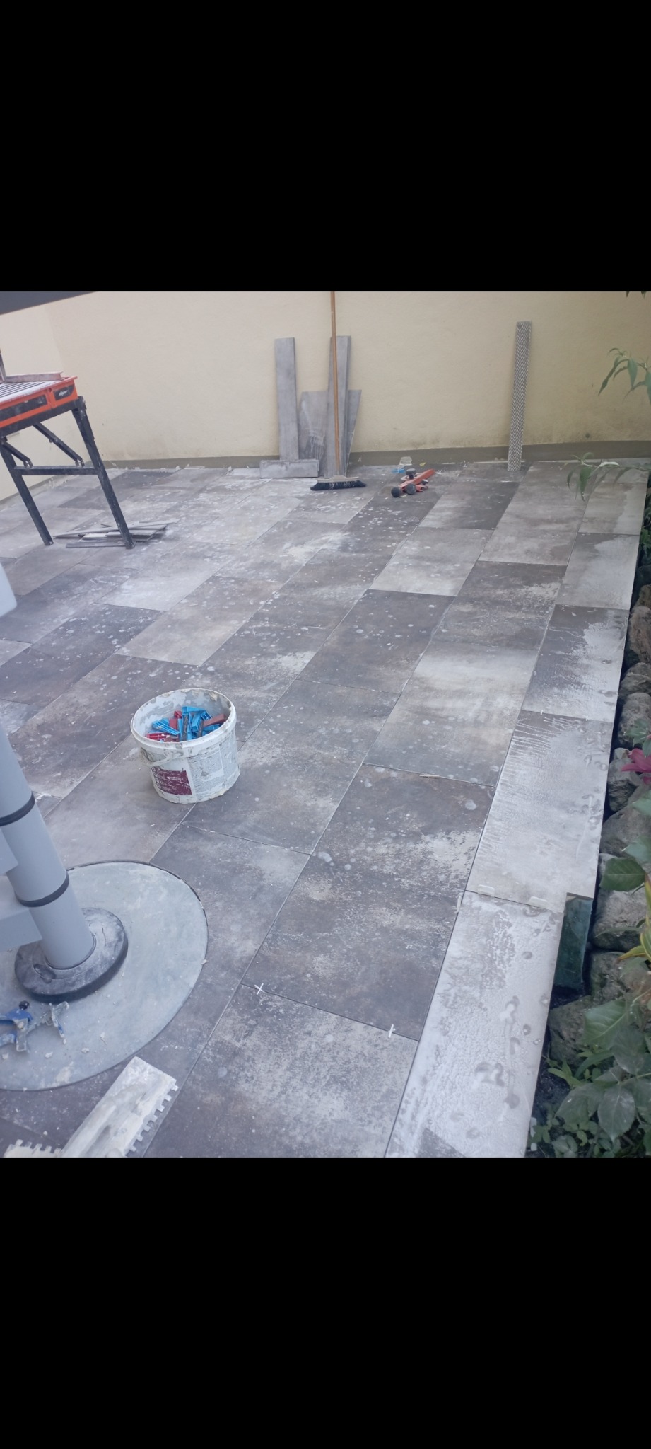 Układanie szarych płytek na zewnątrz, widoczny biały pojemnik z klipsami do płytek, narzędzia glazurnicze i fragment roślinności.