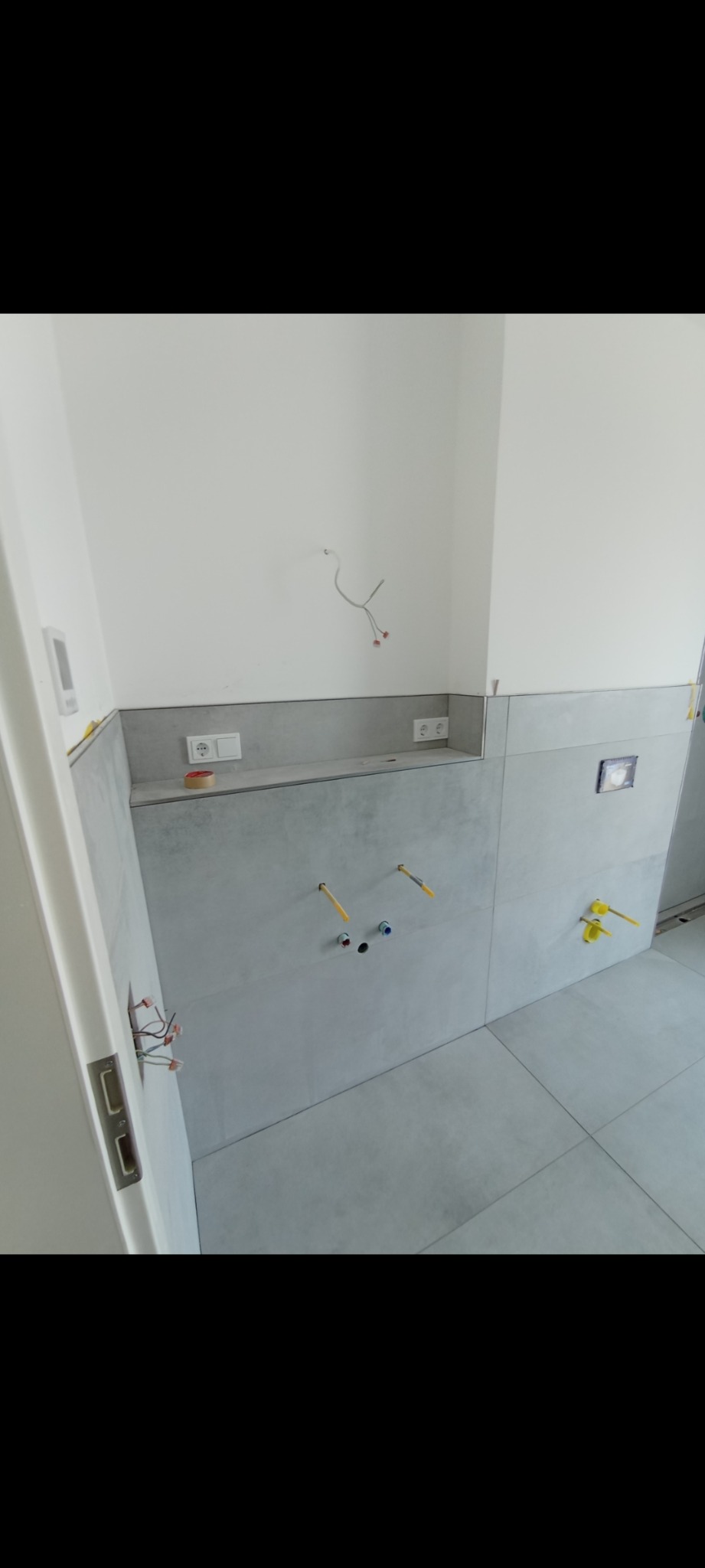 Szare płytki na ścianie i podłodze w trakcie montażu instalacji wodnej, widoczne wystające rurki i kable elektryczne.