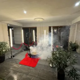 Oświetlona platforma 360 otoczona barierami ze sznura, z efektem dymu i bańkami mydlanymi, ustawiona na zewnątrz budynku z roślinami w donicach i kamienną podłogą.