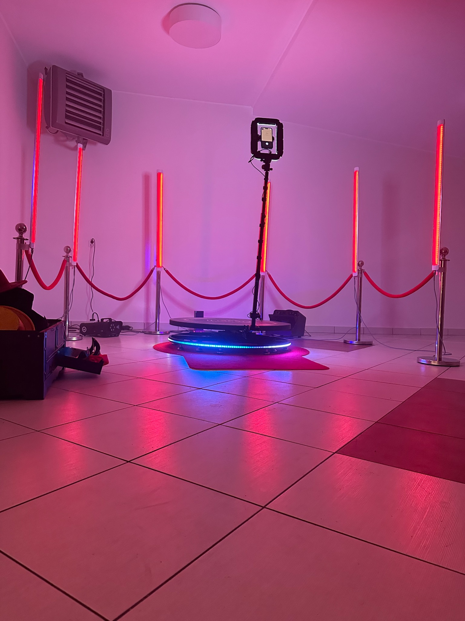 Rotacyjna platforma 360 stopni z oświetleniem LED i uchwytem na smartfon, otoczona słupkami z czerwonym sznurem, ustawiona w pomieszczeniu z kafelkową podłogą i czerwonym dywanikiem.