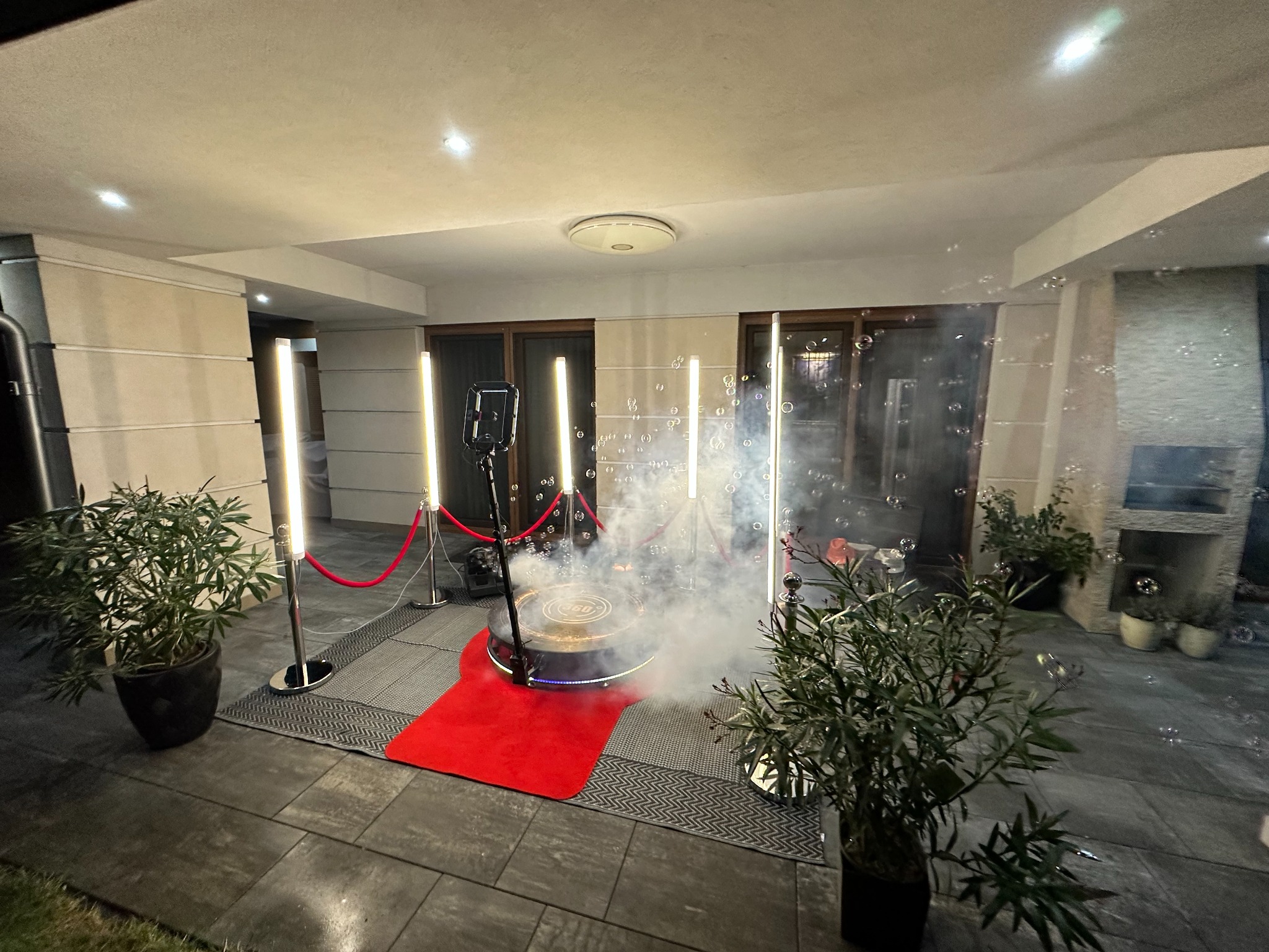 Oświetlona platforma 360 otoczona barierami ze sznura, z efektem dymu i bańkami mydlanymi, ustawiona na zewnątrz budynku z roślinami w donicach i kamienną podłogą.