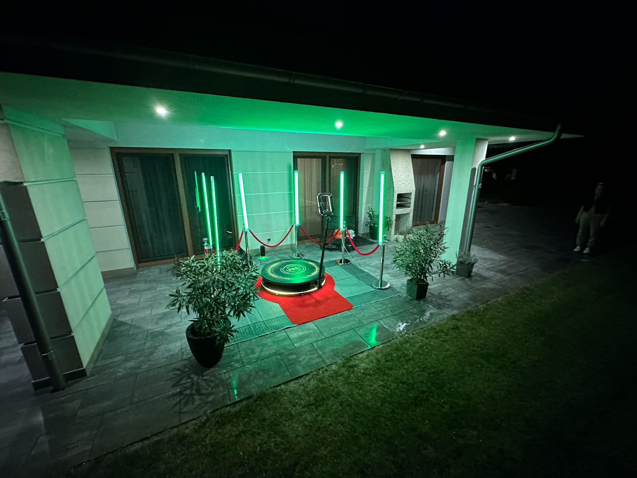 Oświetlona na zielono platforma obrotowa z czerwonym dywanem, otoczona słupkami z czerwonym sznurem i roślinami w doniczkach, ustawiona na zewnątrz budynku w nocy.
