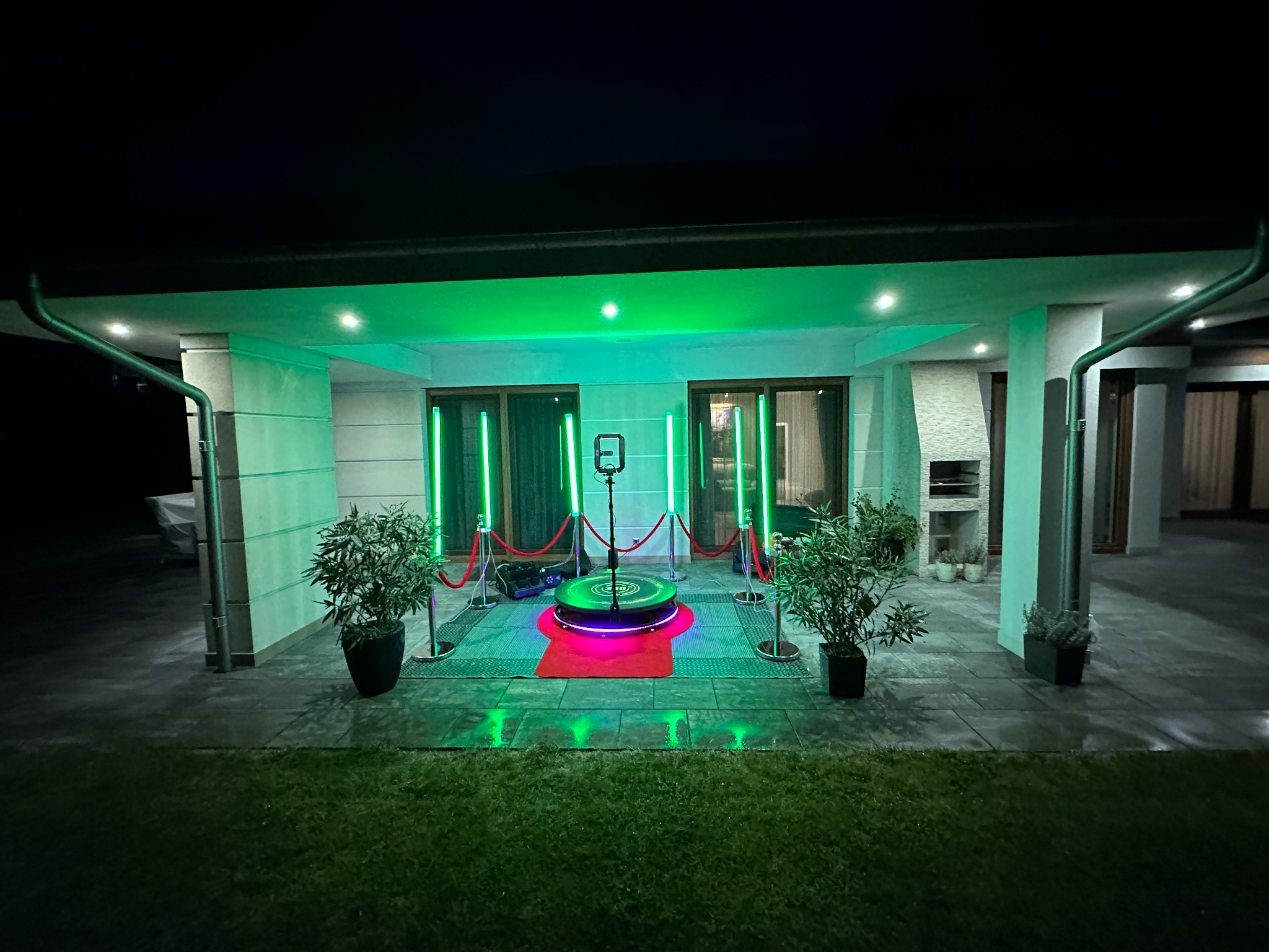 Rozstawiona, oświetlona fotobudka 360 na zewnątrz domu w Płocku, z czerwonym dywanem i roślinami doniczkowymi po bokach, gotowa do użycia.