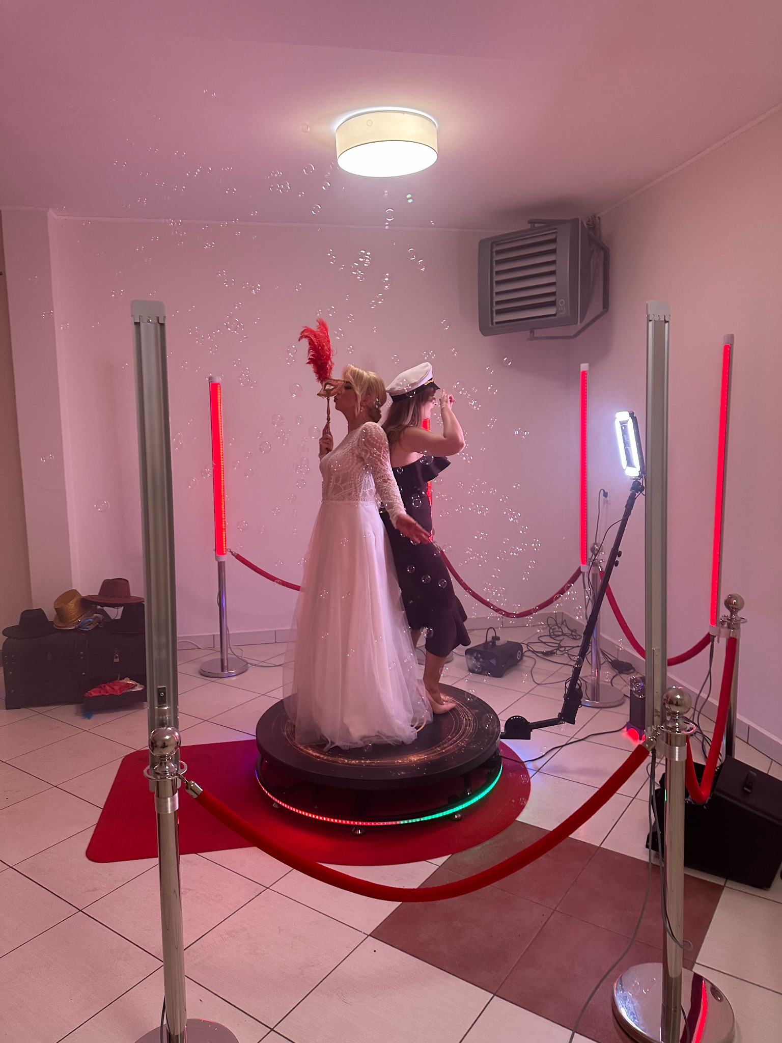 Dwie kobiety, jedna w białej sukni ślubnej z maską z piórami, druga w czarnej sukience i czapce, bawią się na obrotowej platformie otoczonej czerwonymi słupkami i ledowymi lampami, w tle widoczna...