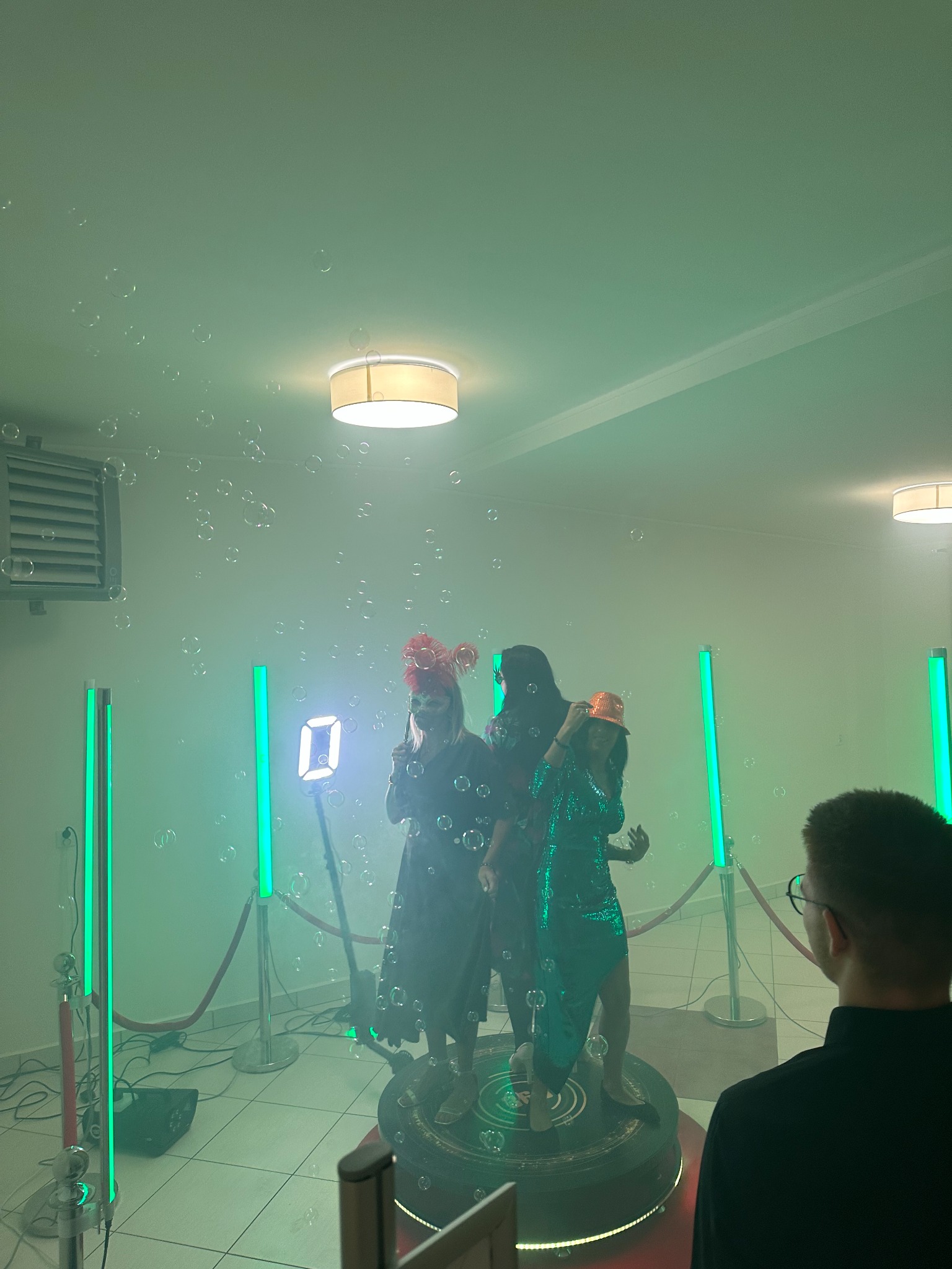 Trzy osoby w kostiumach pozują na platformie otoczonej bańkami mydlanymi i neonowymi lampami w zielonym oświetleniu, widziane z perspektywy obserwatora z tyłu.