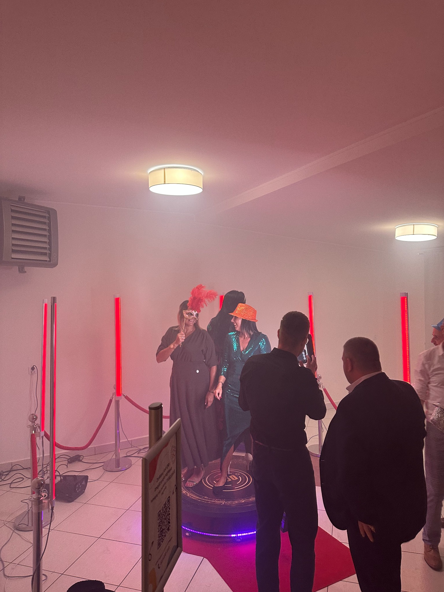 Uczestnicy imprezy pozują w fotobudce 360 stopni z czerwonym oświetleniem, jeden z gości filmuje ich telefonem.