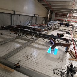 Usługi spawalnicze Jakub Śledź - Spawanie rur metalowych w hali przemysłowej, widoczne spawane elementy, narzędzia i osprzęt spawalniczy, całość na tle konstrukcji stalowej i instalacji pod stropem.