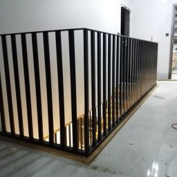Usługi spawalnicze Jakub Śledź - Nowoczesna balustrada schodowa z czarnych pionowych profili i białych paneli, zamontowana na drewnianej podstawie, widok z góry schodów, w tle fragment łazienki.