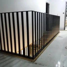 Nowoczesna balustrada schodowa z czarnych pionowych profili i białych paneli, zamontowana na drewnianej podstawie, widok z góry schodów, w tle fragment łazienki.