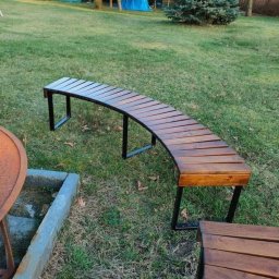 Usługi spawalnicze Jakub Śledź - Zakrzywiona ławka parkowa z drewnianym siedziskiem i metalowymi nogami, ustawiona na trawniku w ogrodzie, z widocznym fragmentem betonowego obrzeża i zarysem namiotu w tle.